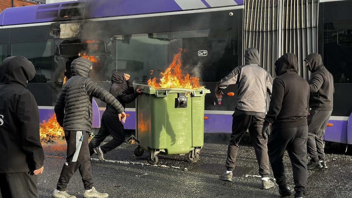 A bus being set on fire by protestors holding a garbage bin on fire in east Belfast