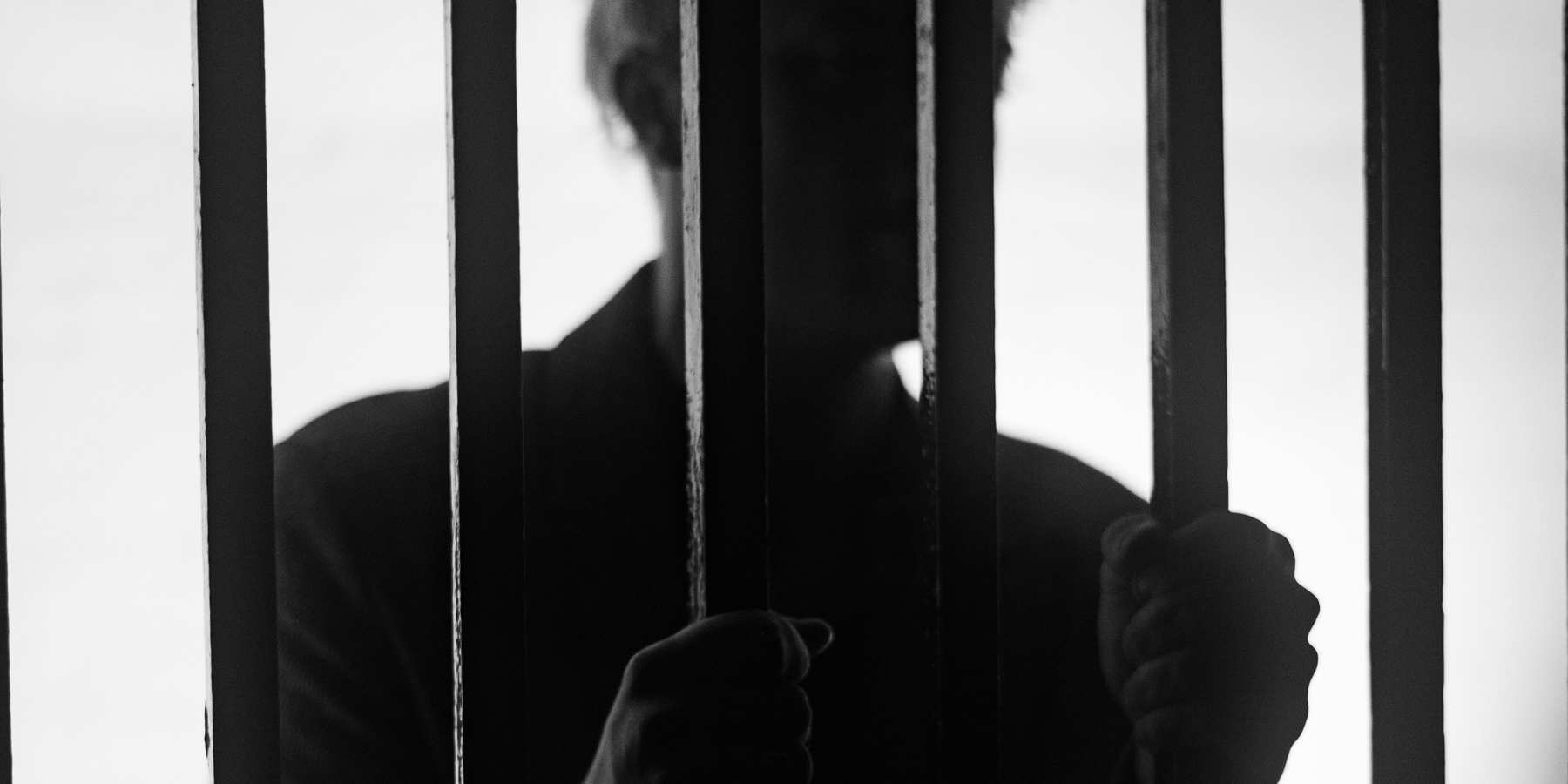 A man standing behind bars in a jail cell silhouetted.