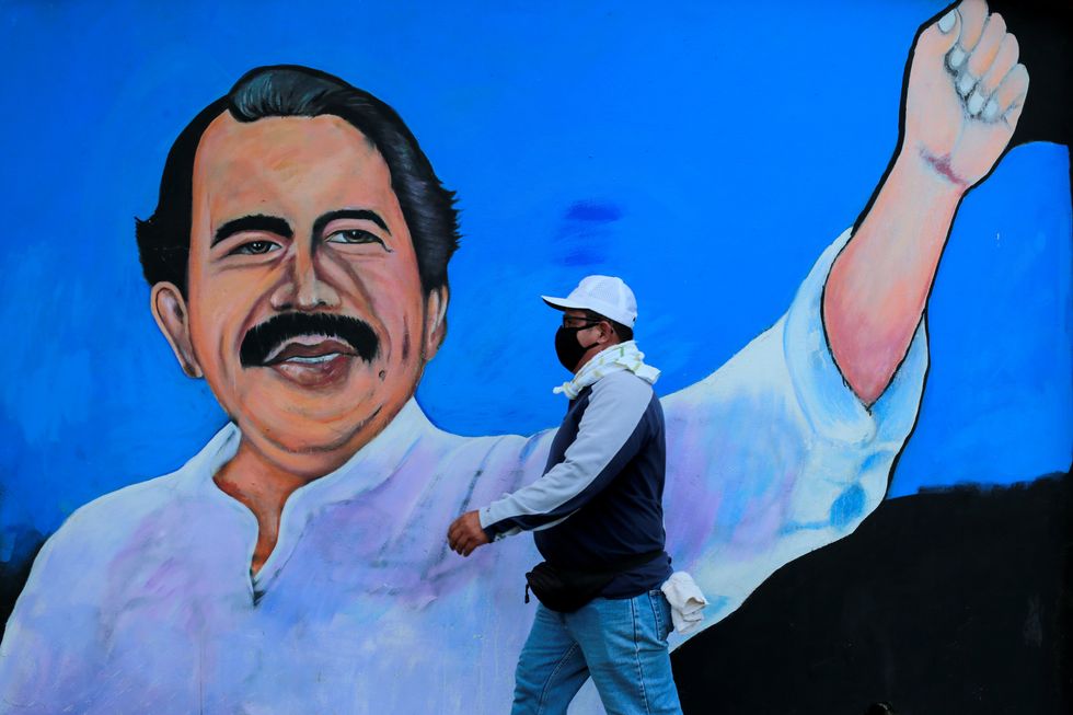 A masked man walking past a painting of Daniel Ortega, President of Nicaragua