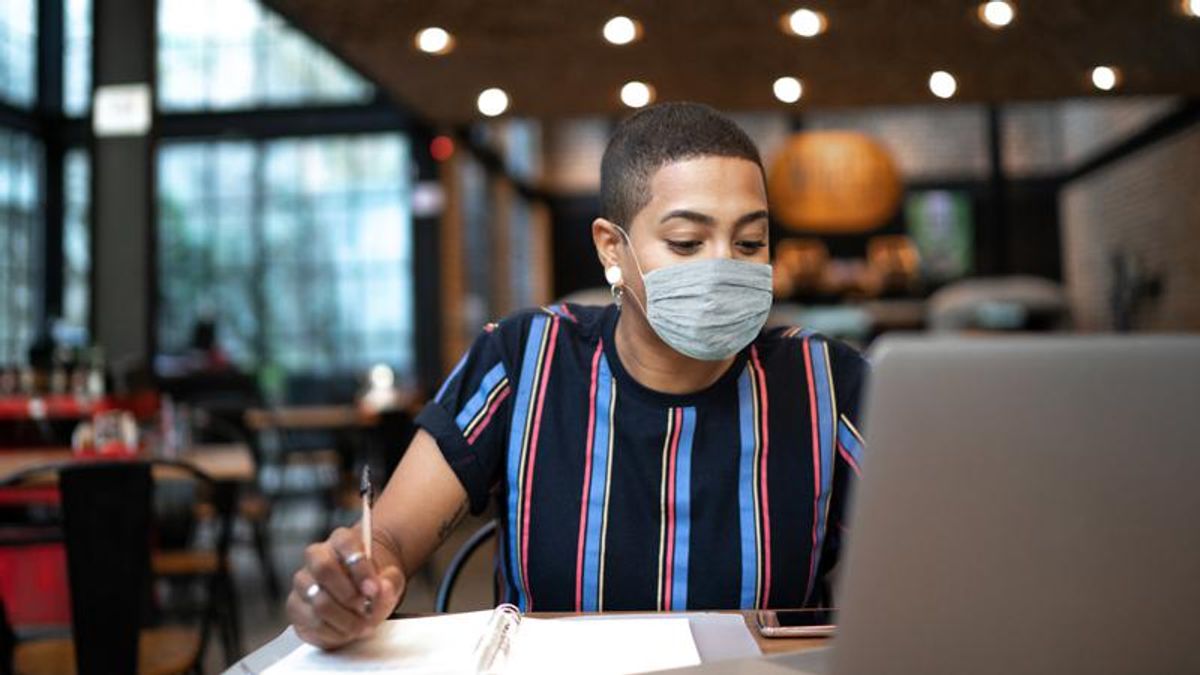 A person of color wearing a cloth mask working at a laptop
