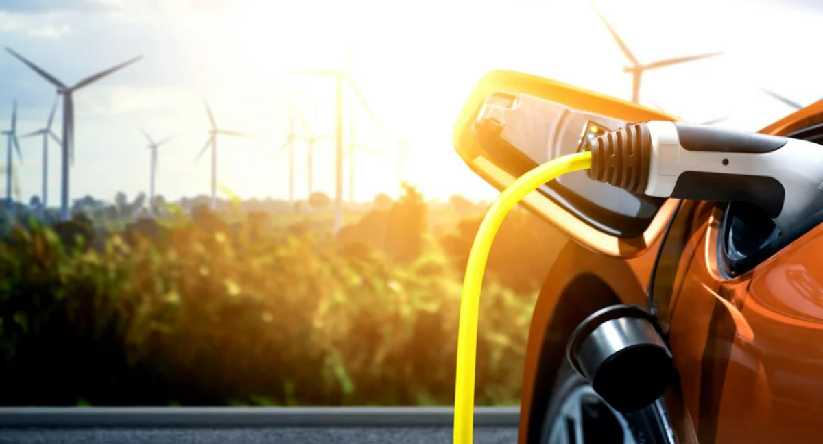 A red car being charged in the foreground with wind turbines in the background