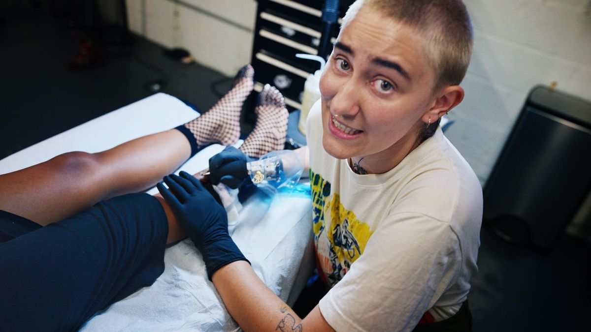 A tattoo artist smiling at the camera while working on a client