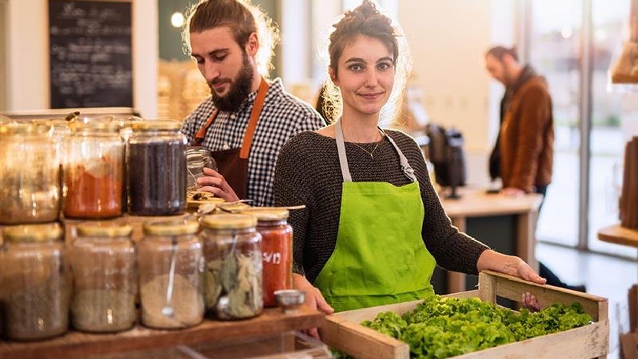 A woman holding a crate of leafy greens and a man behind her taking out spices from a jar