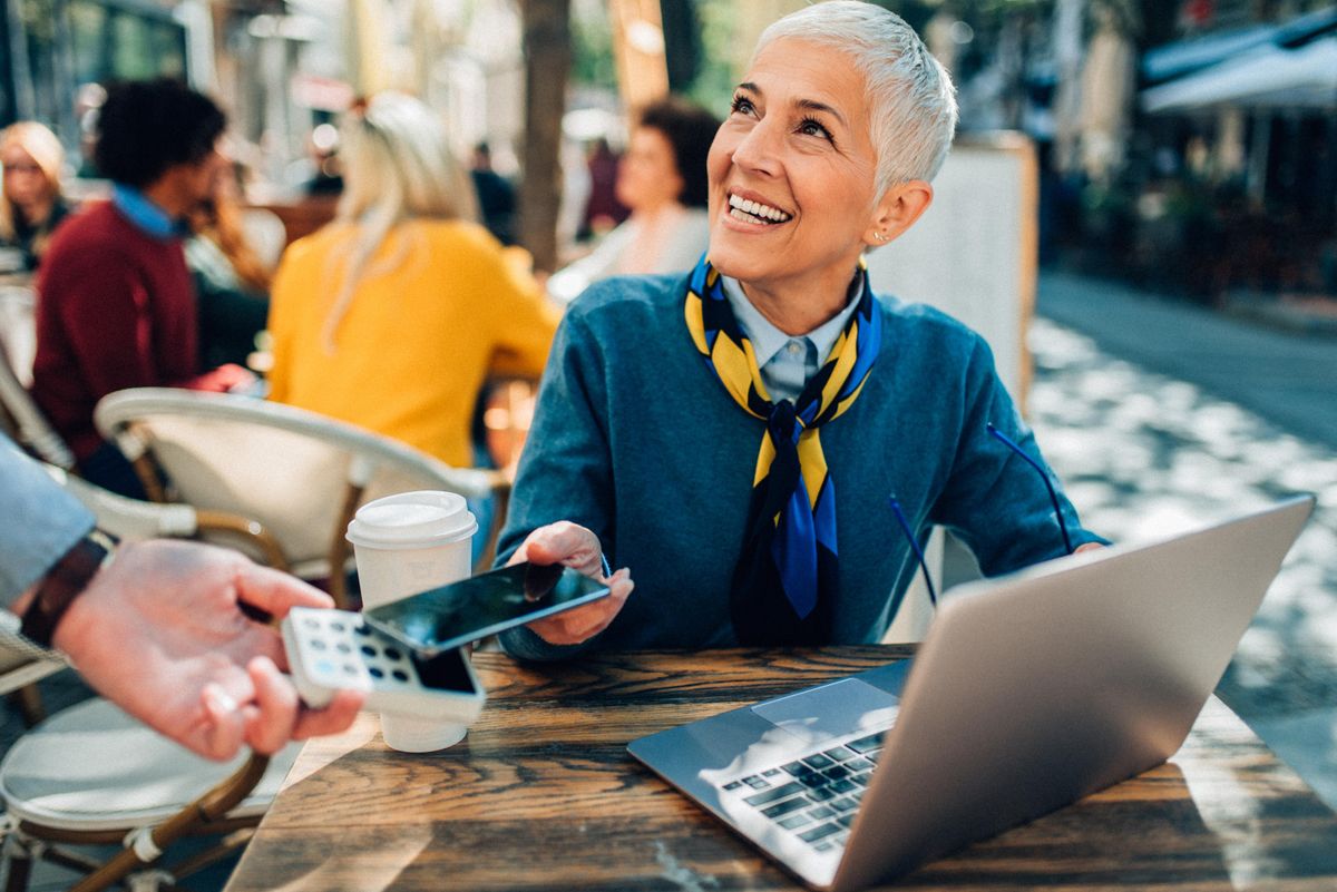 A woman smiling with an open laptop in front of her at a sidewalk cafe reaching out to make a digital payment