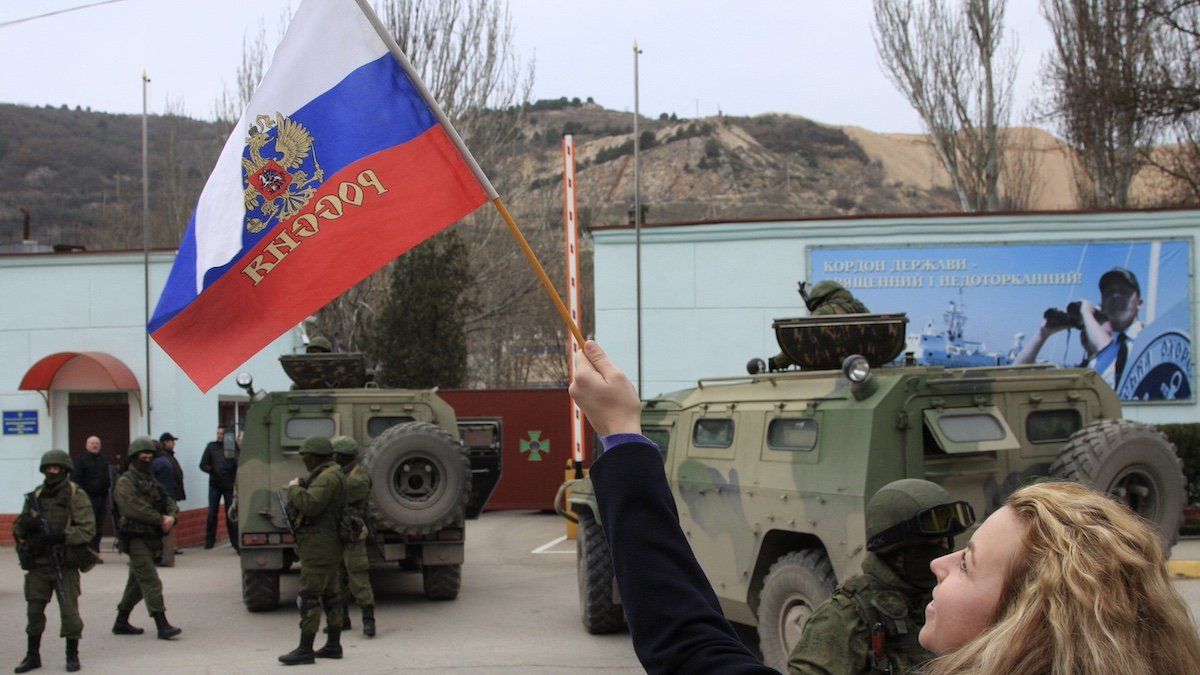 ​A woman waves a Russian flag as armed servicemen wait near Russian military vehicles outside a Ukrainian border guard post in the Crimean town of Balaclava March 1, 2014. Russian President Vladimir Putin wrested control of the Ukrainian Black Sea region of Crimea from Kiev on Saturday citing a threat to Russian citizens and servicemen of the Russian Black Sea fleet based there. 