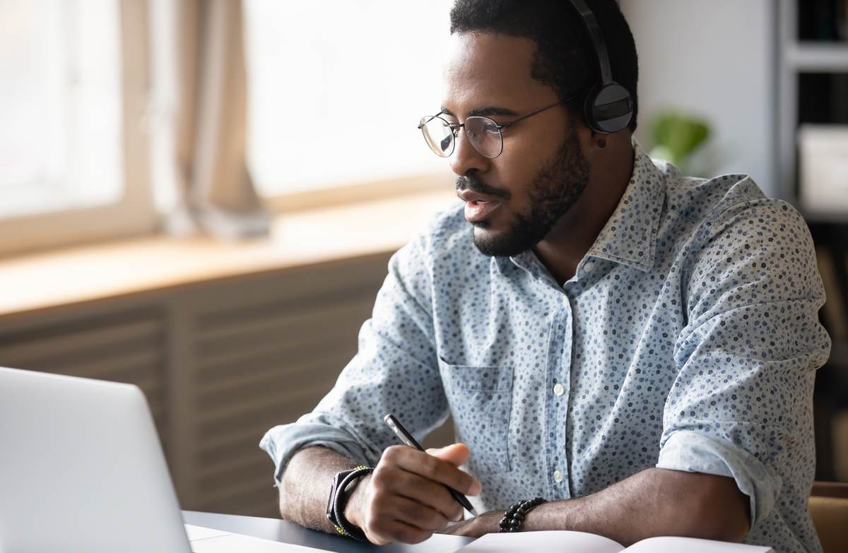 African American man wearing headphones, looking at a laptop with sunlit windows in the background