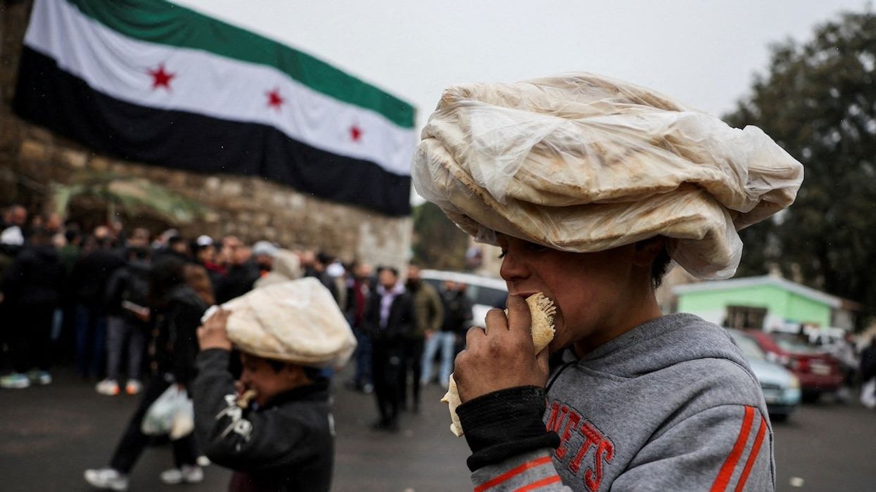 FILE PHOTO: Children eat bread on a street near a flag adopted by the new Syrian rulers, after the ousting of Syria's Bashar al-Assad, in Damascus, Syria, December 24, 2024.