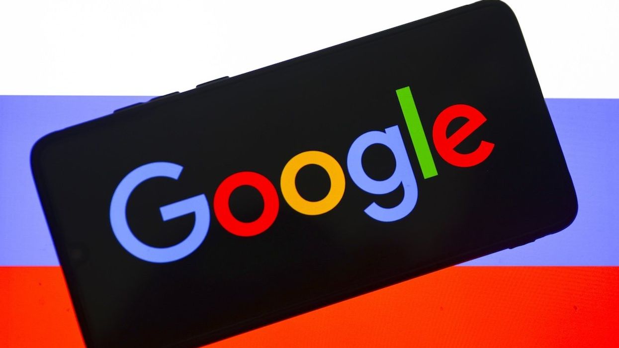 Google logo is displayed on a mobile phone screen photographed with a Russian flag in the background for an illustration.