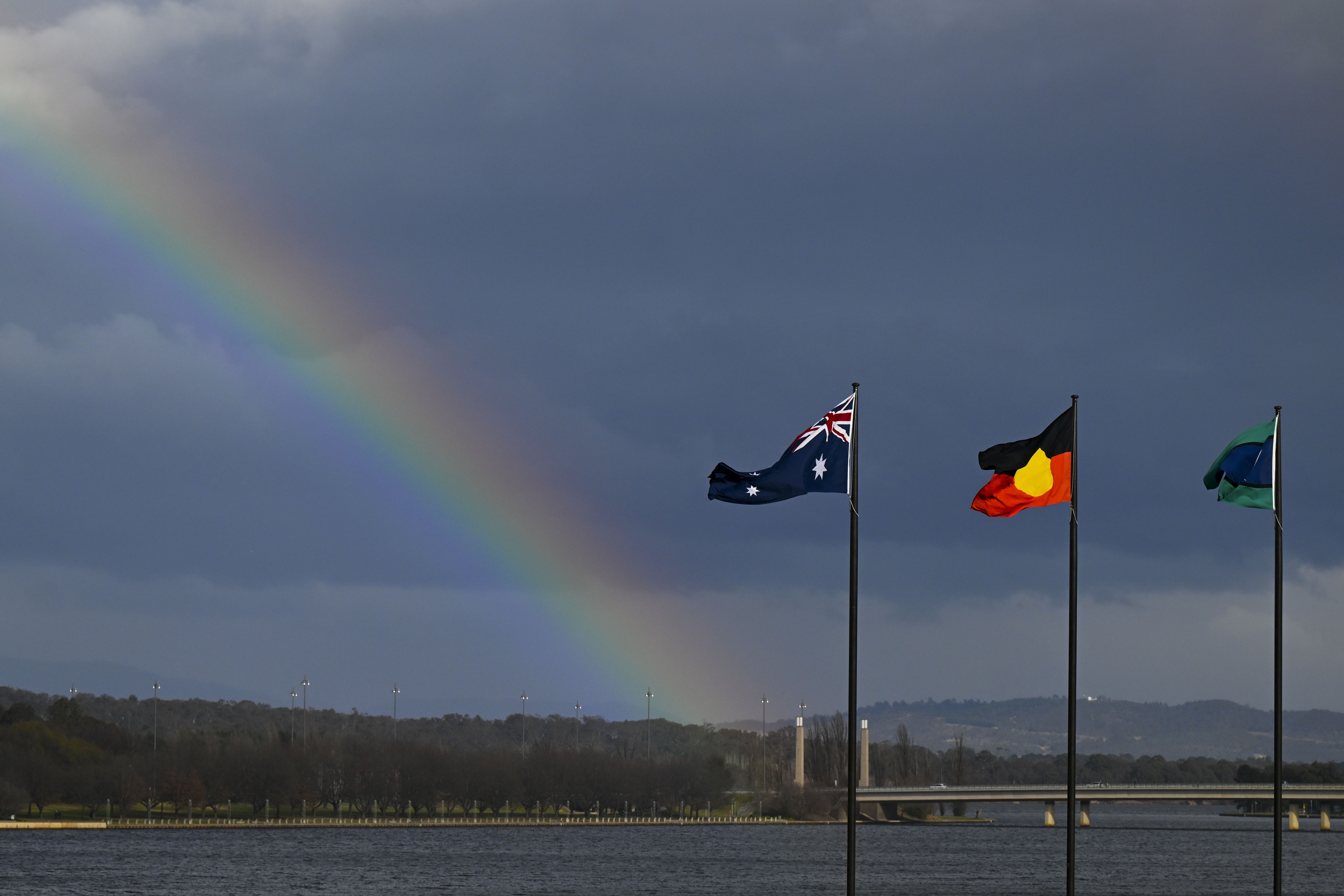 A rainbow is seen behind the Australian flag, the Indigenous flag and the flag of the Torres Strait Islands in Canberra, Friday, July 28, 2023.