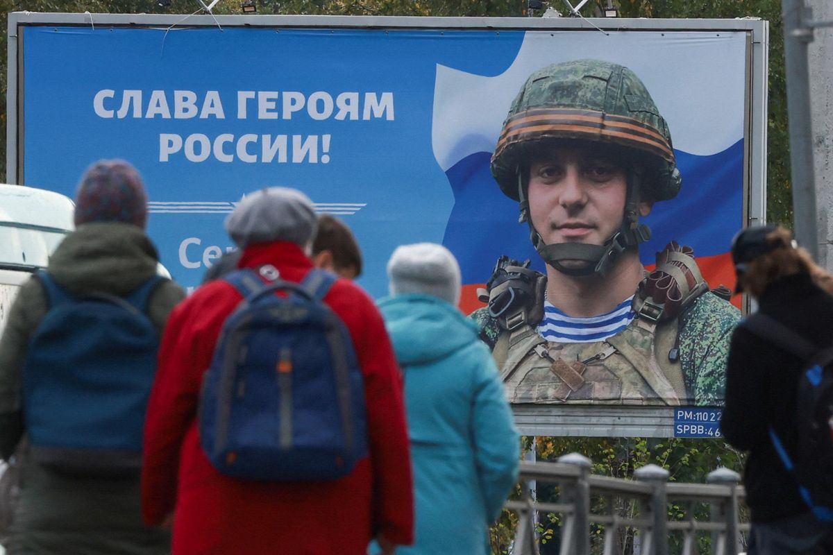 People gather at a tram stop in front of a board displaying a portrait of a fallen Russian soldier in St. Petersburg. A slogan reads: "Glory to heroes of Russia!" 