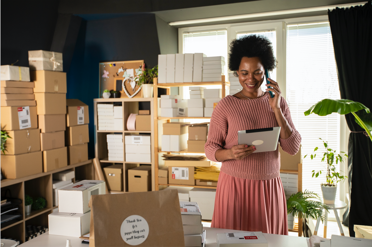 Photograph of a small business owner, a smiling Black woman in a pink knit sweater and skirt, looking at a tablet and surrounded by boxes
