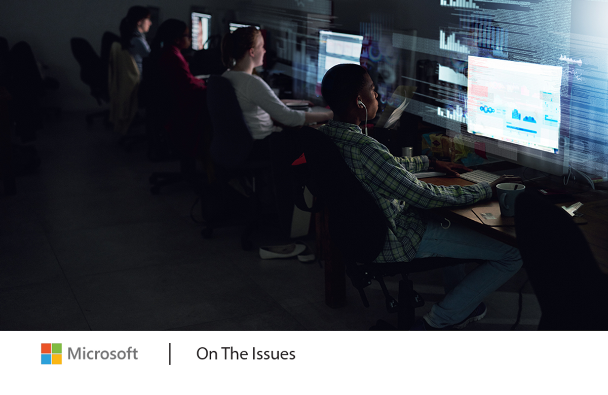 Several students sitting and working in a darkened room with computer screens brightly lit in front of them