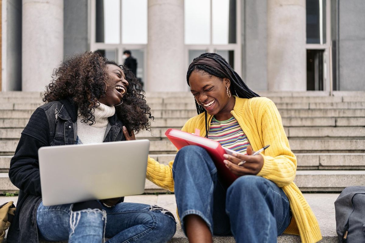 Two African American women appear laughing while sitting on the steps of a building. One with a laptop; the other with a binder and pen.