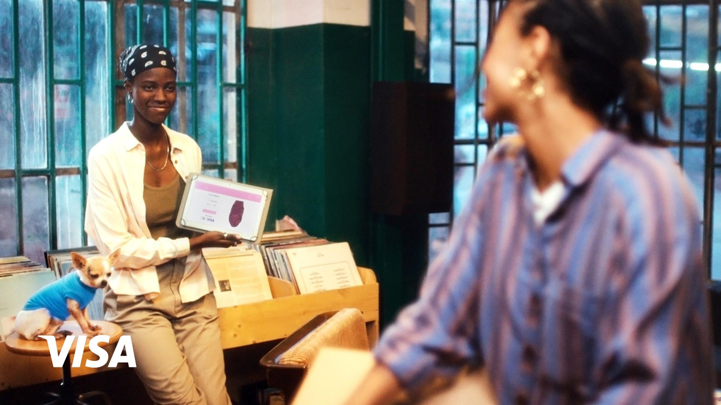 Two Black women looking at one another across a record store, one showing a screen displaying an online Visa purchase