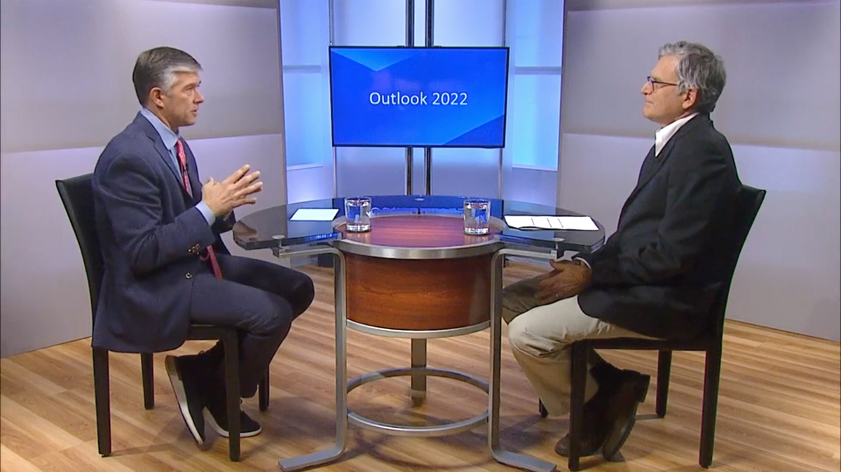 Two men facing one another in dialogue with Outlook 2022 appearing on a screen between them