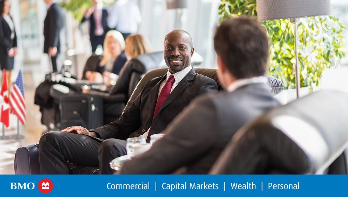 Two men wearing suits sit in lobby armchairs for a meeting.