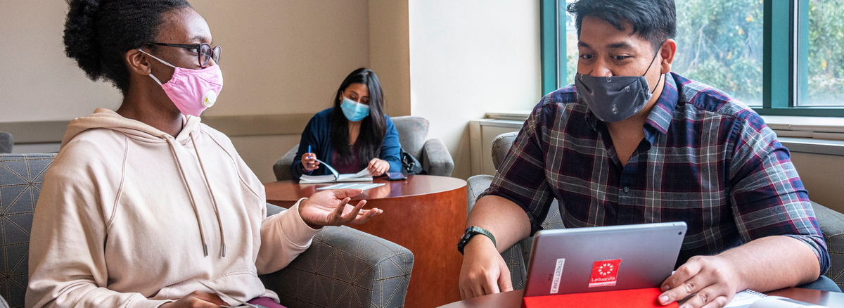 Two people in foreground, wearing masks, appear to be working together at a tablet computer; A woman in background also wearing a mask