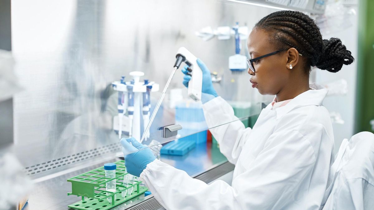 Woman of color wearing a white coat and blue gloves working in a lab.