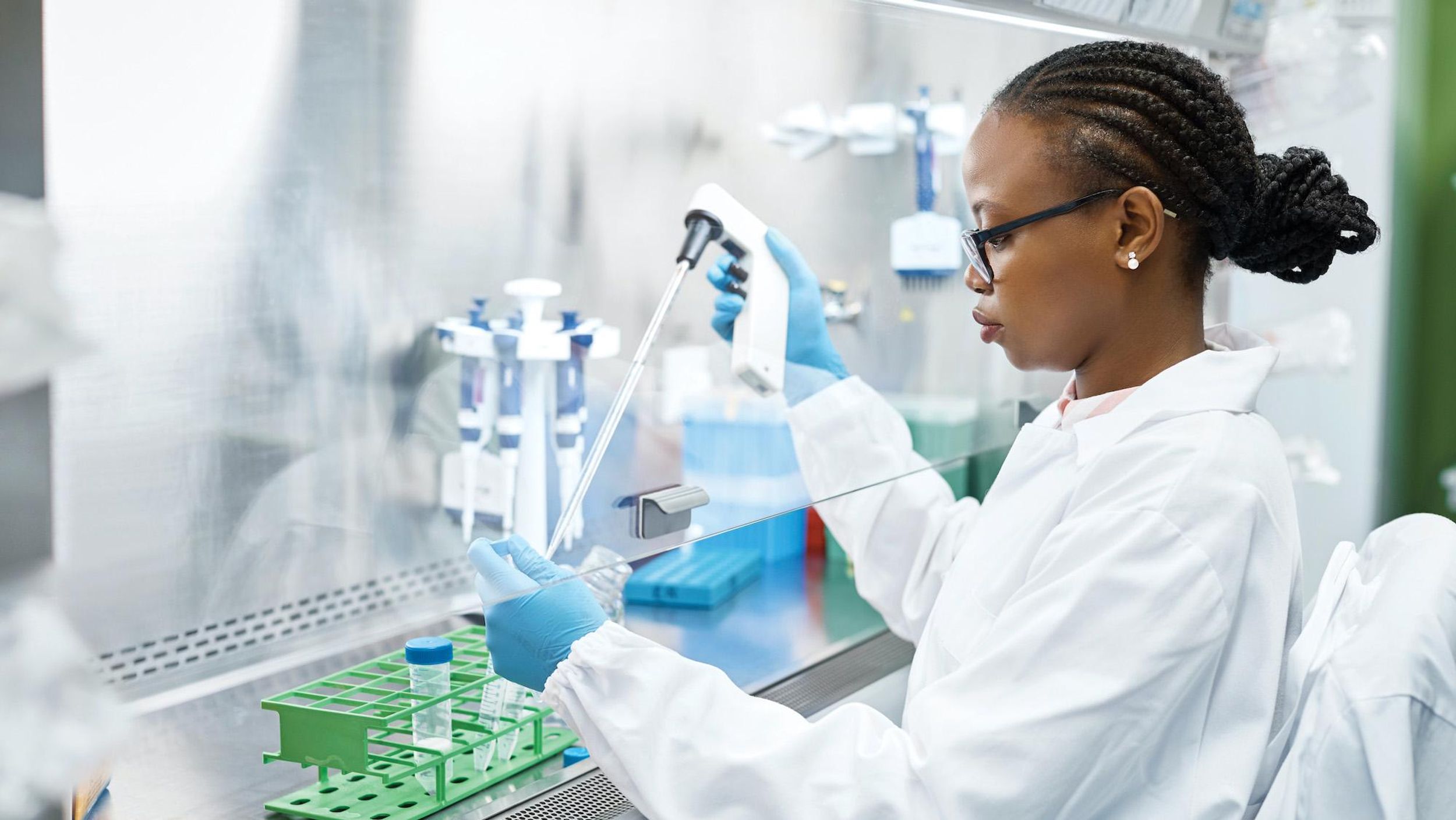 Woman of color wearing a white coat and blue gloves working in a lab. 