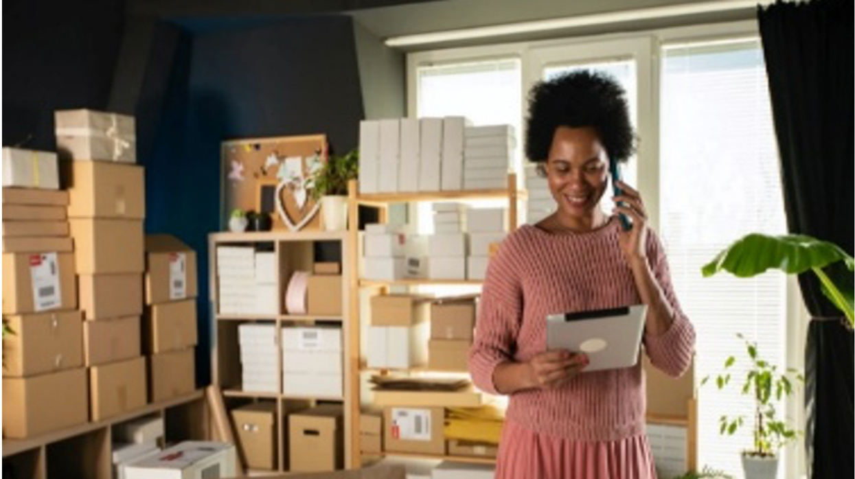 Woman smiling on the phone, while she holds her tablet in a living room full of boxes and plants.
