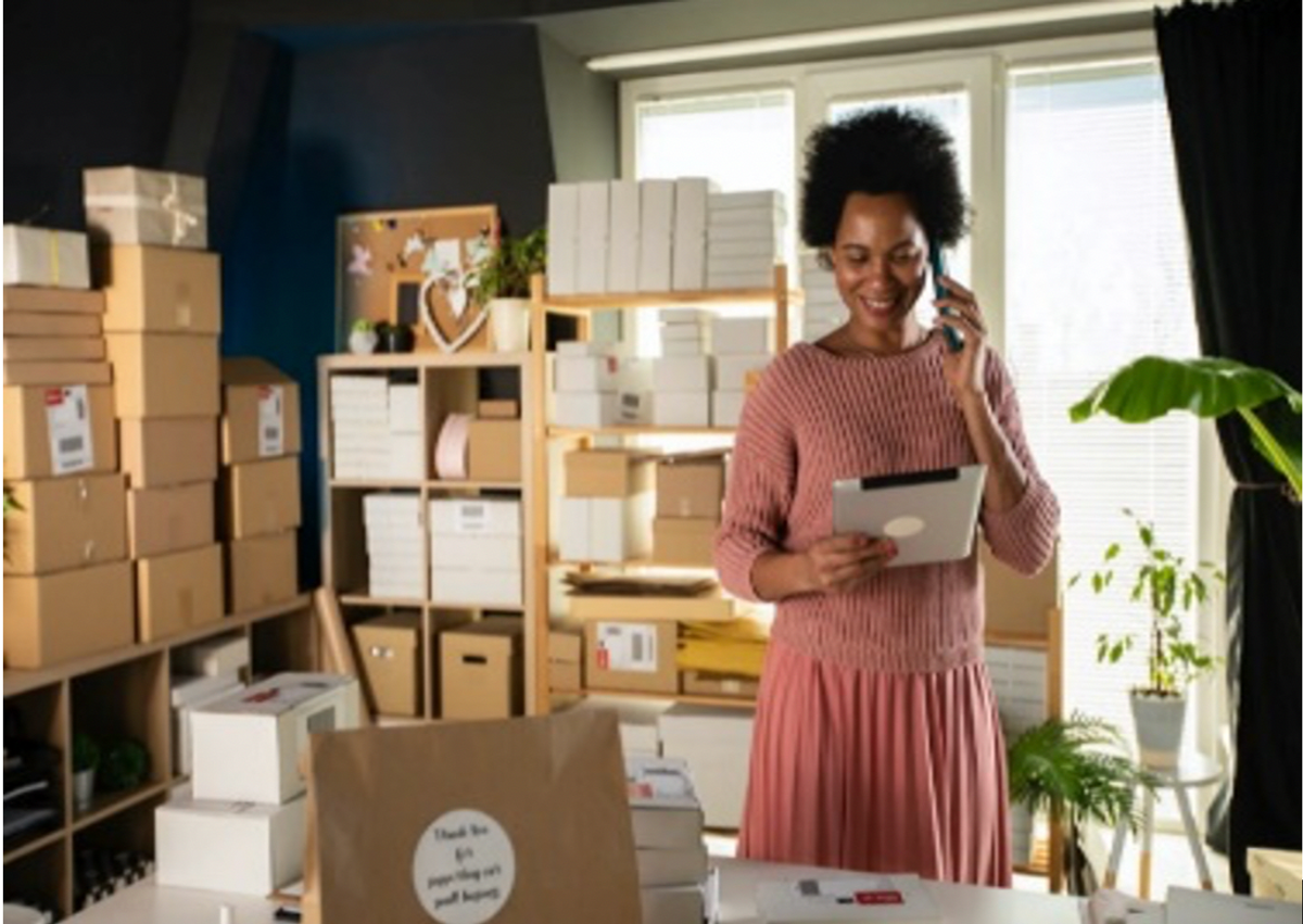 Woman smiling on the phone, while she holds her tablet in a living room full of boxes and plants.