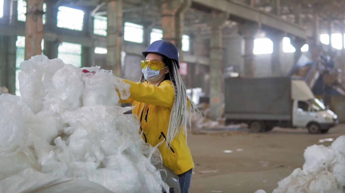 Woman wearing a yellow coat, sunglasses, a hat, and a mask recycling.